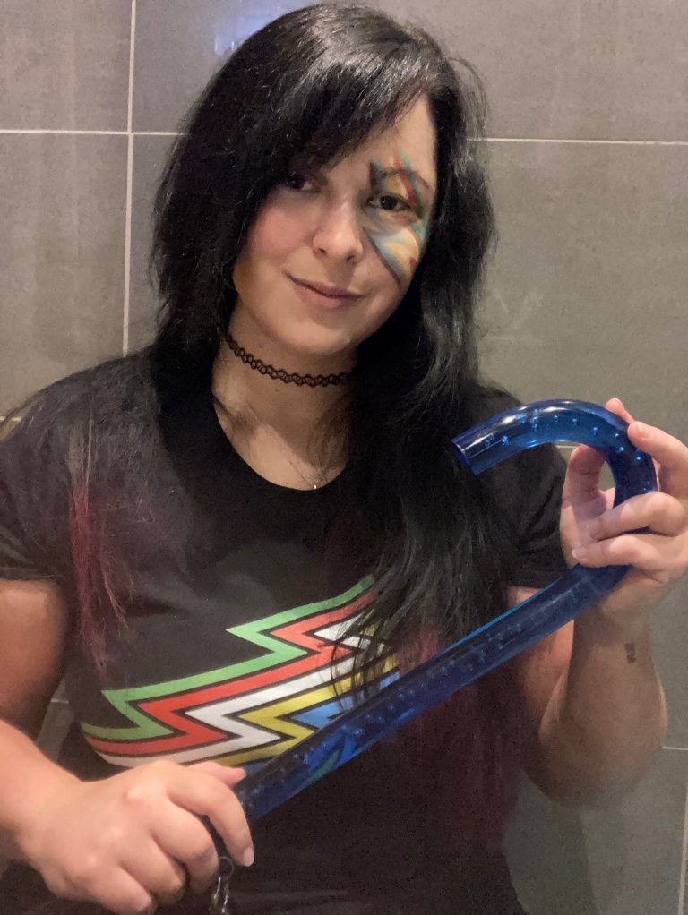 Ruthy is a white woman with long black hair. She is wearing a black t-shirt with the disability flag on it and has drawn the flag on her face in make-up. She is smiling and holding a blue walking stick.